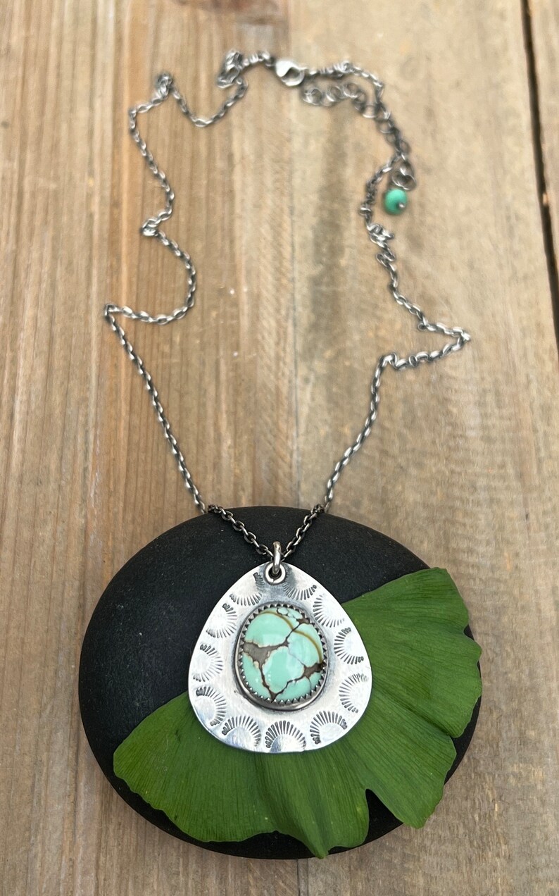 May include: A silver necklace with a teardrop-shaped pendant featuring a green turquoise stone set in a silver frame. The pendant is surrounded by a decorative silver design. The necklace is on a black stone with a green leaf.