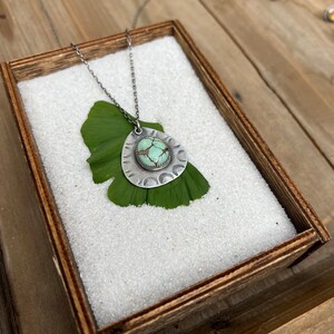 May include: A silver necklace with a turquoise stone pendant. The pendant is surrounded by a decorative silver design. The necklace is displayed on a wooden box filled with white sand.