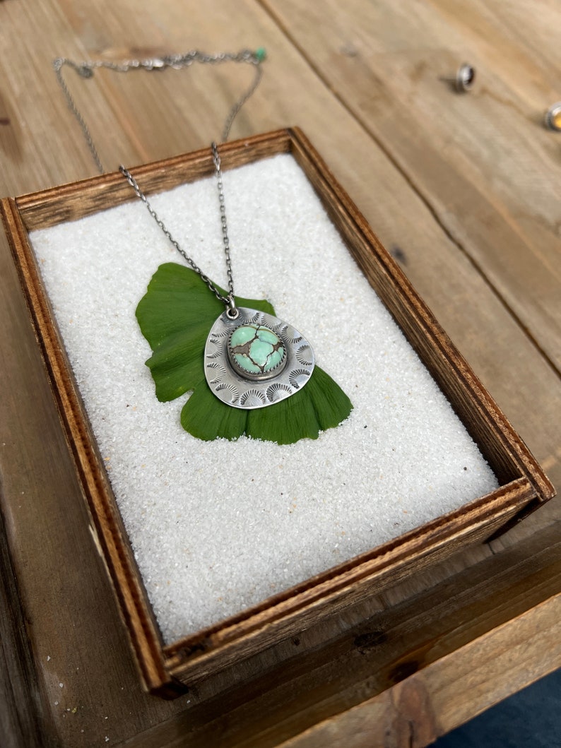 May include: A silver necklace with a turquoise stone pendant. The pendant is surrounded by a decorative silver design. The necklace is displayed on a wooden box filled with white sand.