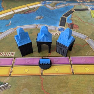 Puede incluir: Un juego de mesa con un mapa y varios edificios en miniatura azules y negros. El tablero del juego tiene un diseño colorido con secciones amarillas, rosas y verdes. Los edificios en miniatura tienen tejados azules y bases negras.