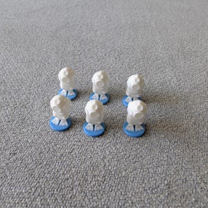 Könnte beinhalten: Sechs weiße, abstrakt geformte Spielsteine mit abgerundeter Oberseite und einem gesichtsähnlichen Design, jeweils auf einem kleinen, runden, hellblauen Sockel montiert. Die Teile sind auf einer grau strukturierten Oberfläche angeordnet.