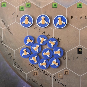 Puede incluir: Fichas de juego azules con diseños naranjas y blancos, dispersas sobre un mapa de juego de mesa. Las fichas presentan un diseño estilizado de nave espacial o avión. El mapa del juego de mesa tiene un esquema de color marrón y beige con espacios hexagonales.