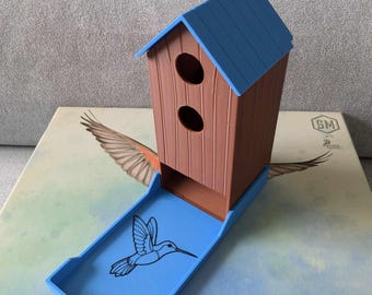 May include: A blue and brown wooden birdhouse shaped dice tower with a hummingbird design on the tray. The tower has two holes on the front and a hinged lid that opens to reveal the tray.