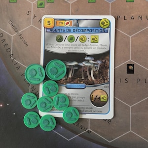 Peut inclure: Une carte de jeu intitulée "AGENTS DE DÉCOMPOSITION" avec une illustration de champignons, accompagnée de plusieurs jetons de jeu verts. La carte présente des symboles et du texte de jeu, avec une valeur de 5 et un indicateur de 3%. Les jetons sont circulaires avec un motif texturé.