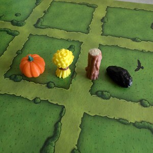 May include: A green game board with four game pieces: a yellow sheaf of wheat, a brown log, an orange pumpkin, and a black rock.