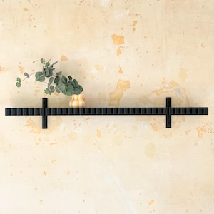 May include: A long, black, horizontally mounted wall shelf with a repeating fluted or ribbed texture along its front edge. The shelf is supported by two black vertical brackets and holds a small, yellow and white striped vase with eucalyptus.