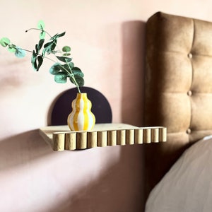 May include: A wooden shelf with a scalloped edge, mounted on a wall. A small yellow and white striped vase with green leaves sits on the shelf.
