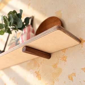 May include: A wall-mounted shelf featuring a light-coloured plywood surface with visible layered edges and a dark brown cylindrical wooden support bracket. The shelf holds a small pink and white striped vase and a green leafy plant.