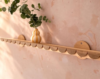 May include: A long wooden wall shelf featuring a scalloped front edge and two circular mounting brackets. A small, bulbous ceramic vase with vertical yellow and white stripes sits on the shelf, containing a green leafy plant branch.