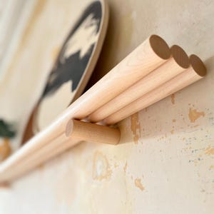 May include: A wooden wall shelf featuring three cylindrical dowels and a long shelf. The shelf is crafted from light-coloured wood and is mounted on a wall with a distressed, textured finish. A decorative round object with a black and white design rests on the shelf.