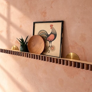 May include: A long, horizontal wall shelf featuring a fluted dark wood front edge and two semi-circular brass mounting brackets. On the shelf sits a small green potted succulent, a round wooden bowl, and a framed print of a rooster.