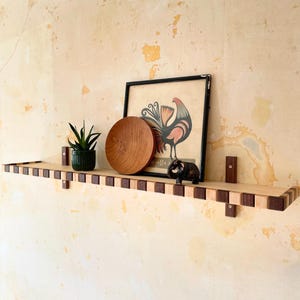 May include: A wooden shelf with a decorative edge, displaying a framed rooster print, a wooden bowl, a small potted plant, and a figurine. The shelf is mounted on a textured, light-coloured wall. The shelf's edge has alternating light and dark wood blocks.