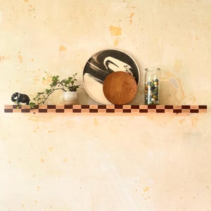 May include: A wooden shelf with a checkerboard pattern holds decorative items. A black and white plate, a small wooden plate, a glass jar with marbles, a small potted plant, and a figurine are displayed on the shelf.