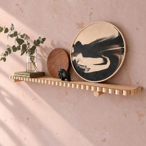 May include: A light-coloured wooden wall shelf with a scalloped front edge. On the shelf sits a clear glass vase with green eucalyptus stems, a small dark brown wooden circular disc, a black elephant figurine, and a large round tray with black and beige abstract paint patterns.