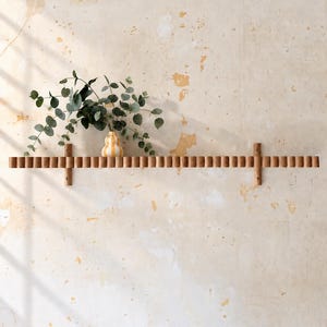 May include: A long, horizontal wooden wall shelf featuring a repeating fluted or ribbed texture along its front edge. The shelf is mounted with two vertical wooden supports and holds a small, yellow and white striped vase with green eucalyptus.