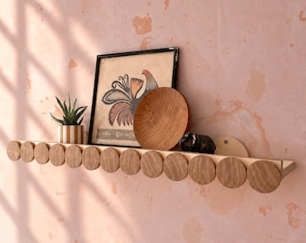 May include: A long, horizontal wooden wall shelf featuring a front edge decorated with a series of uniform, circular wooden discs. The shelf is mounted on a textured, light pink wall and holds a small potted succulent, a framed illustration, a wooden bowl, and a small dark figurine.