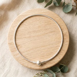 May include: A silver-toned snake chain necklace featuring a smooth, round bead clasp at the centre. The necklace is formed into a circular shape, showcasing the flexible, textured metal links of the chain and the polished finish of the spherical closure.