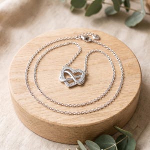 May include: A silver-toned necklace featuring a heart-shaped pendant intertwined with an infinity symbol. The pendant is embellished with small, clear, sparkling stones along the heart outline. The chain is a delicate, thin, silver-toned link style.