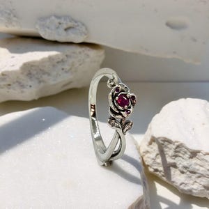 May include: A silver ring with a rose design, featuring a central, round, red gemstone. The rose is adorned with smaller red gems and detailed leaves. The ring is set against a white backdrop.