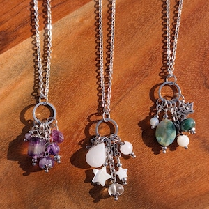 May include: Three silver necklaces with different charm clusters. One has purple beads, another has white teardrop and star charms, and the third features green and white stones with a lotus charm. The necklaces hang against a wooden background.