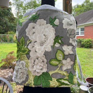 Puede incluir: Una bolsa de cordón gris con un estampado floral blanco y verde. La bolsa está hecha de tela de lino y tiene un cordón blanco.