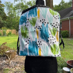 Può includere: Un marsupio per neonati in tessuto bianco con un bordo a pois neri e una stampa astratta colorata con colori blu, verde, giallo e bianco.