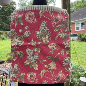 Può includere: Un grembiule rosso a fiori con un bordo a righe verde e bianco. Il grembiule presenta un motivo floreale con rose e foglie.