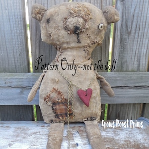 Puede incluir: Un oso de trapo hecho a mano con un colgante en forma de corazón y un solo ojo de botón. La muñeca está hecha de tela de arpillera y tiene un aspecto desgastado. El texto "Pattern Only--not the doll" está escrito en la imagen. El texto "Crows Roost Prims" también está escrito en la imagen.