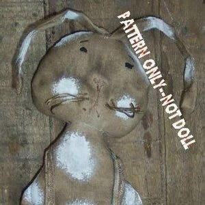 May include: A handmade primitive style bunny doll with white painted accents. The doll is wearing overalls and holding a small floral patterned pillow. The doll is made from burlap fabric and has a rustic look. "PATTERN ONLY - NOT DOLL" is written on the image.