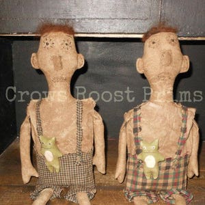 May include: Two primitive-style dolls with tan fabric bodies, brown hair, and stitched facial features. They wear plaid overalls with a frog applique. The text "PATTERN ONLY - NOT THE DOLL" is at the bottom.