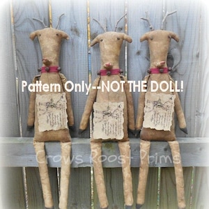 May include: Three handmade primitive style reindeer dolls with brown fabric bodies, red ribbon collars, and brown paper accents. The dolls are sitting on a wooden fence. The text "Pattern Only - NOT THE DOLL!" is printed on the image.