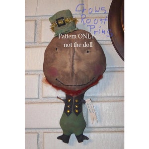 Puede incluir: Una muñeca de fieltro marrón con un sombrero verde y un borde dorado. La muñeca lleva un atuendo verde y negro con botones dorados. La muñeca sostiene un cartel que dice "On the luck of the Irish". El texto "Crows Roost Prims Pattern ONLY not the doll" está escrito encima de la muñeca.