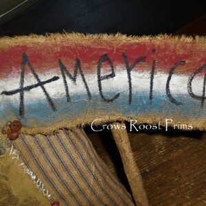 May include: A rustic sign with the word "America" painted in black on a burlap banner. The banner features red, white, and blue stripes, with frayed edges and small bell accents. The sign is part of a larger decorative piece.