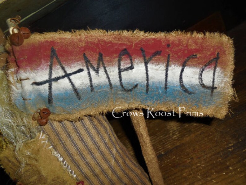 May include: A rustic sign with the word "America" painted in black on a burlap banner. The banner features red, white, and blue stripes, with frayed edges and small bell accents. The sign is part of a larger decorative piece.