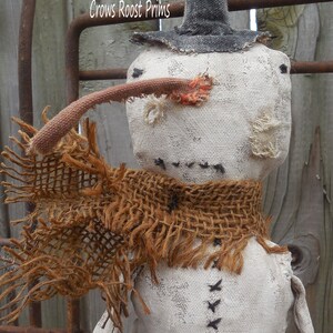 May include: A white fabric snowman with a black top hat, a brown burlap scarf, and a red and orange fabric nose. The snowman is standing in front of a wooden fence.