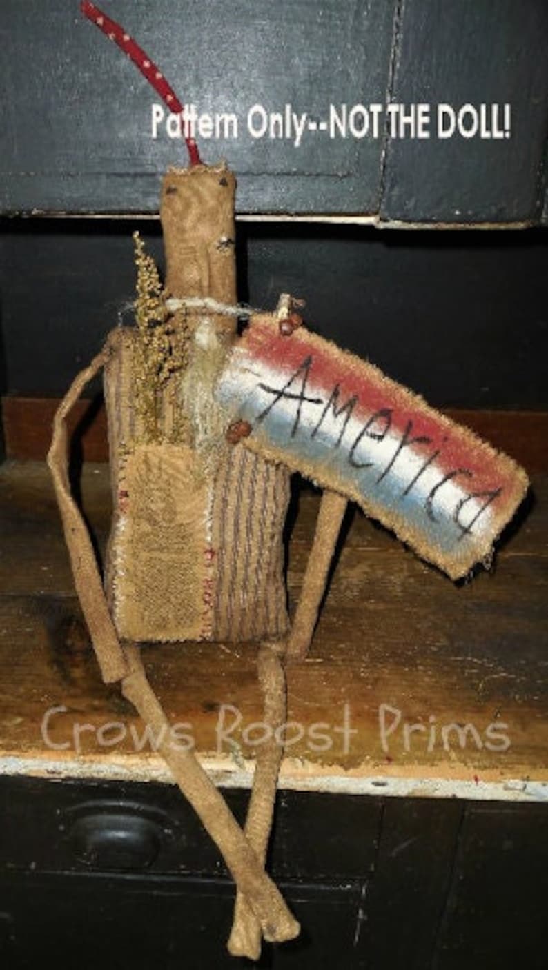 May include: A rustic doll with a burlap body, wooden limbs, and a sign that reads "America." The sign is painted with red, white, and blue stripes. The doll has a twig-like head and arms, and is decorated with dried foliage.
