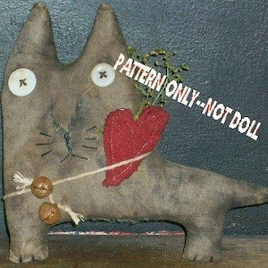 May include: A handmade, stuffed cat doll with button eyes, whiskers, and a red heart. The cat is tan with darker markings and has two small bells tied around its neck. The text "PATTERN ONLY--NOT DOLL" is printed across the image.