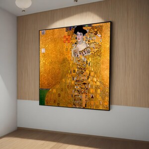 Può includere: Stampa incorniciata di un dipinto di Gustav Klimt. L'opera raffigura una donna con un abito dorato e un cappello nero su uno sfondo dorato con motivi dettagliati. Il dipinto è in una cornice nera.