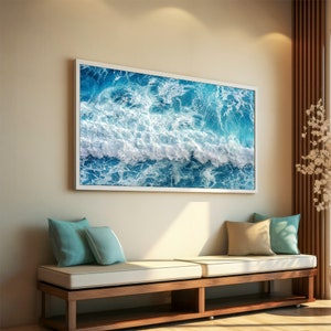 May include: A framed photograph of a blue ocean with white waves crashing. The photograph is hanging on a beige wall above a wooden bench with white cushions and blue throw pillows.