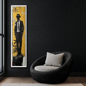 May include: A black and white silhouette of a person wearing a hat and suit standing in front of a yellow and white background. The image is framed and hanging on a black wall next to a gray wicker chair with a gray cushion.