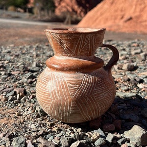 Könnte beinhalten: Ein brauner Tontopf mit einem weißen geometrischen Muster. Der Topf hat eine breite Öffnung und einen Henkel an einer Seite. Er steht auf einem Bett aus kleinen Steinen.