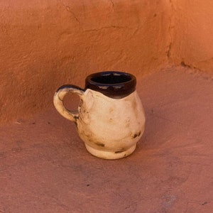 Peut inclure: Une petite tasse en céramique blanche avec un bord et une anse marrons. La tasse est posée sur une surface orange texturée.