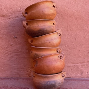 Könnte beinhalten: Ein Stapel aus sechs Terrakotta-Schalen mit abgerundeten Kanten und kleinen Griffen. Die Schalen sind in einer vertikalen Säule gestapelt, wobei die größte Schale unten und die kleinste oben liegt. Die Schalen sind hellbraun und haben eine leicht raue Textur.