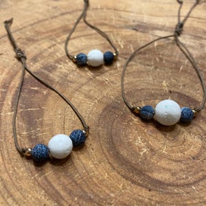 Könnte beinhalten: Drei verstellbare Armbänder an braunen Kordeln, jedes mit einer zentralen weißen, porösen Perle, flankiert von zwei blauen Perlen mit Rissmuster und kleinen metallisch goldfarbenen Zwischenelementen. Die Armbänder sind gruppiert angeordnet.