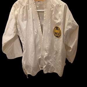 May include: A white martial arts gi jacket with long sleeves and a V-neck collar. The garment features a circular black and yellow patch on the left chest displaying a fist icon and Japanese characters. A tan label with the text JSK is at the neck.