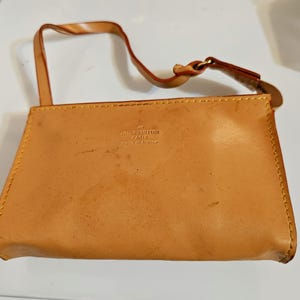 May include: A tan leather clutch with a short strap. The clutch has a rectangular shape with rounded corners and a zipper closure. The words "LOUIS VUITTON PARIS made in france" are printed on the front.