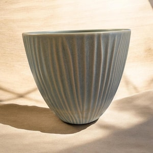 May include: A ceramic bowl with a soft blue-grey glaze and a vertical ribbed texture. The bowl features a wide, open circular rim that tapers down to a smaller base, showcasing a consistent pattern of raised lines around the entire exterior surface.