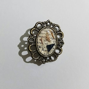 May include: A bronze oval-shaped brooch with a decorative filigree design. The center of the brooch features a clear resin oval with a piece of paper inside. The paper has text printed on it and a small seashell and a black triangle are embedded in the resin.