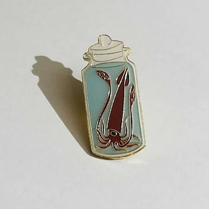 Op de afbeelding: Een gouden emaille pin met daarop een inktvis in een glazen pot. De inktvis is rood en heeft een wit oog. De pot is gevuld met blauwe vloeistof.