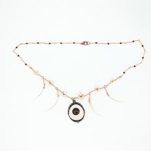 May include: A delicate gold necklace with a white eye pendant and dangling bone charms. The necklace features small red beads and a lobster clasp closure.